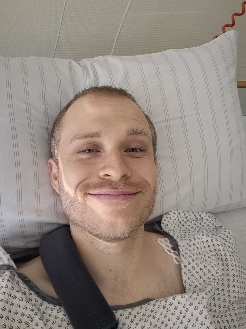 Michi lächelnd im Krankenhausbett, den linken Arm in einer Schlinge